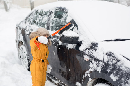 A small five-year-old Child brushing snow off car after storm. Kid with winter brush and scraper...