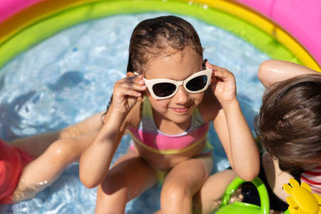 A wet girl in sunglasses is sitting in a small inflatable pool with clear water on a hot sunny day. Using a protective baby sunscreen. Children have fun in the water.の写真素材