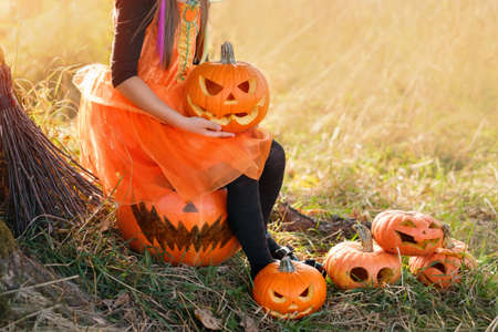 A girl in an orange carnival dress holds a pumpkin with an evil smile carved in her hands. The Halloween holiday. Jack-o - lantern. a few pumpkins are standing on the grass in the sun. Without a faceの写真素材