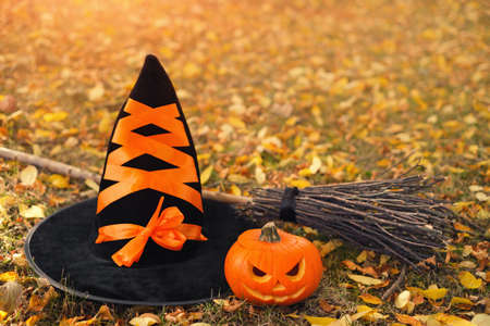 A witch's black hat with an orange ribbon next to a broom and a pumpkin with a sinister face carved on the ground among the fallen autumn dry leaves. Halloween holidayの写真素材