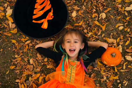 A girl in a witch costume lies in the autumn leaves, next to a pumpkin and a hat, with her mouth open. Trick or treating. Jack o 'lantern. Autumn seasonal discounts and sales for Halloweenの写真素材