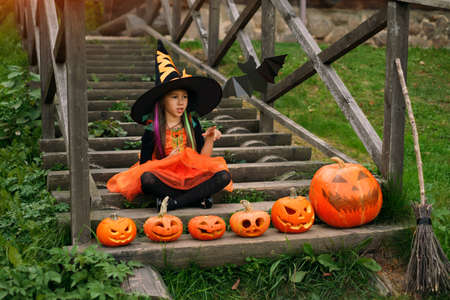 A girl dressed up as a witch pointing with thumb finger aside at copy space. Pumpkins stand on the steps of the ladder. Traditions of decorating the yard in front of the house on Halloween.の写真素材