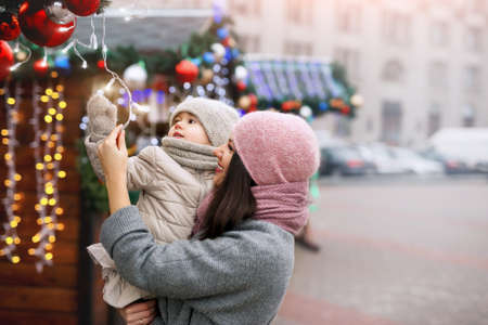 A mother and her little daughter walk through the city streets and Christmas markets decorated with garlands with lights, fir branches and Christmas balls. A cold frosty day on New Year's Eveの写真素材
