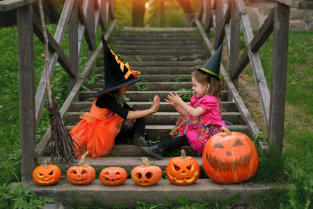 Two sisters in carnival costumes of witches are sitting on an old staircase and playing with their palms. The decor is made of carved Jack o Lantern pumpkins. Halloween. Trick or treat. steps at houseの写真素材