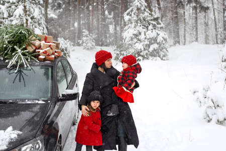 A mother with two daughters in the forest next to a car, on the roof of which there is a Christmas tree with gifts. A snowy frosty day on the eve of New Year and Christmasの写真素材