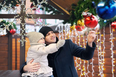 A father and daughter walk through the city streets and markets decorated with garlands with lights, fir branches and Christmas balls. A girl in knitted mittens stretches out her hand to the ballの写真素材
