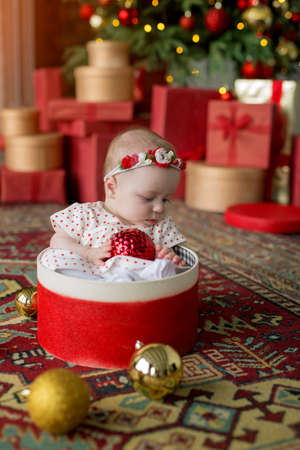 A little girl in a white dress with red polka dots is sitting in a gift round box with a Christmas ball in her hands against the background of a Christmas tree garland with unfocused lightsの写真素材