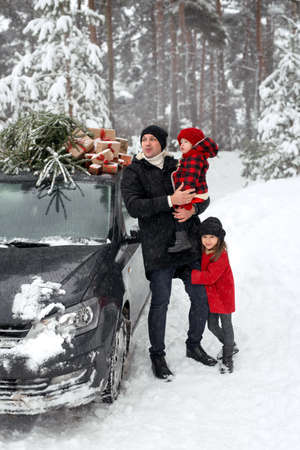 A father and daughters at the car with a Christmas tree and gifts look away with a surprised face. Daughter in dad's arms on a cold snowy winter day. Christmas fairs and discounts.の写真素材