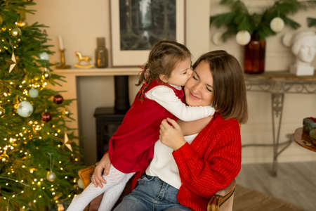 A little daughter kisses and hugs her mom on Christmas Eve. A child and a mother in a room with a Christmas tree on the eve of the new year. Family holidayの写真素材