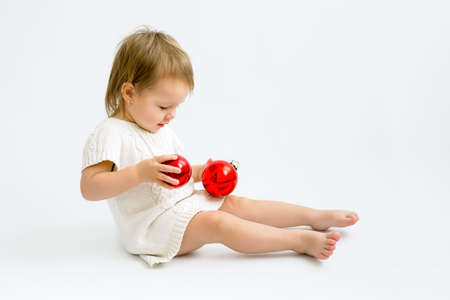 A child in a knitted sweater is playing with two red glass Christmas balls. A girl on a white background with space to copy. Waiting for the New Year. One - year -old kidの写真素材