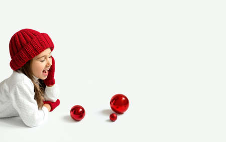 A girl in a red hat and mittens shouts aside at copy space, putting her palm to her mouth. Three Christmas glass balls in front of the kid. Children are waiting for Christmas and New Year. Bannerの写真素材