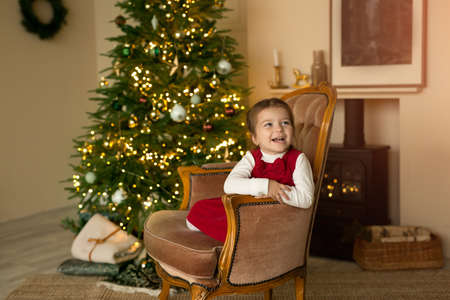 A cute cheerful laughing child is sitting in an armchair by the fireplace against the background of a Christmas tree with lights. Cozy room with Christmas decorationsの写真素材