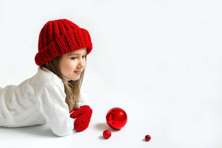 Pensive dreamy girl in a knitted red winter hat with Christmas balls on a white studio background with a place to copy. Looking forward to the New Year and Christmas.の写真素材
