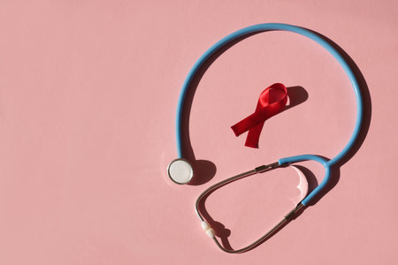 Red ribbon loop and stethoscope on pink studio background with deep shadow. World awareness AIDS Day. Campaign to combat HIV infection. Copy space. A symbol of hope and protest against discriminationの写真素材