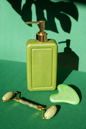 Rectangular green bottle with a jade roller massager and gua sha scraper for face massage on a background with the shadow of a monstera leaf. Skin care and prevention of aging. Modern toolsの写真素材