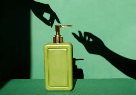 A cosmetic product in a stylish green marble bottle with a golden dispenser and the shadow of elegant female hands on a studio background. Hand skin care with moisturizer and natural soap.Shades greenの写真素材