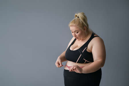 Young full-figured woman with a dazed face measures the size of belly with a measuring tape. Funny blonde obese woman in a sporty black suit, isolated on a gray background. Diet gives a positive resultの写真素材