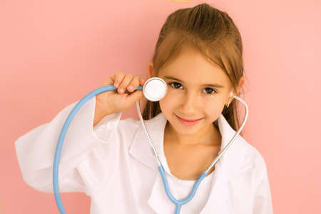 A cute beautiful girl with a stethoscope in her hands and a medical gown on a pink background. Kid portrays a doctor. Children's games in the profession. Pediatric medicine conceptsの写真素材