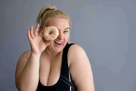 A plump, fat, obese blonde woman in sports clothes closes one eye with a donut, looking through a hole from a bagel. Diet, sports and proper nutrition are the key to a healthy body. Lady shows tongueの写真素材