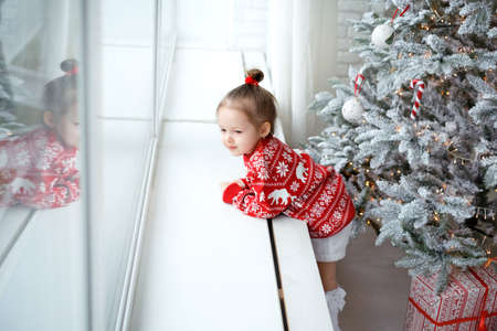 Lovely two-year-old girl is standing at the window thinking and dreaming on the eve of Christmas next to the Christmas tree. Baby in a knitted red sweater with a Christmas ornament with deerの写真素材