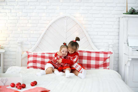 likable gentle girls hug each other tightly while sitting in bed with a gift box. The girl gave her sister a gift and both are happy and laughing. It is joyful to give and receive gifts. Christmasの写真素材