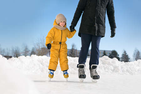 A father teaches a child to skate at a rural outdoor skating rink, holding her hand. The child likes skating. A girl in a winter yellow jumpsuit with frozen rosy cheeks in the cold. Sunny dayの写真素材