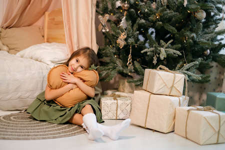 Cute beautiful long-haired brunette girl is sitting on the floor by the bed and the Christmas tree hugging a pillow in the shape of a heart, smiling sweetly. xmas decor of the children's room.New yearの写真素材