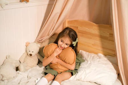 A cute long-haired girl is sitting in a cozy baby bed and hugging a heart-shaped pillow. Comfortable cozy bed and mattress with teddy bears. Children's roomの写真素材