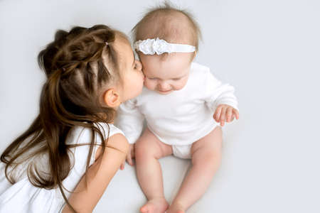 A cute three-year-old girl with pigtails kisses little sister gently on the cheek. Upbringing and mutual understanding in the family. Idyll in the relationship between childrenの写真素材
