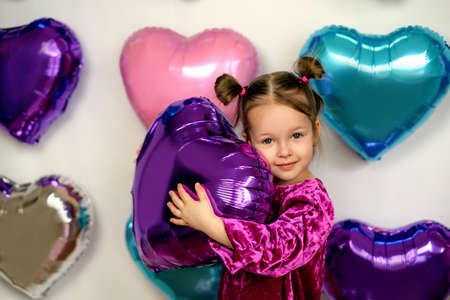 A cute three-year-old girl hugs a purple foil balloon in the shape of a heart. Decor photo zone of hearts for Valentine's Dayの写真素材