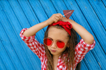 A girl with a red bow on her head and sunglasses holds a triangular shaped lollipop in hands. The child looks into the camera. Blue old painted Background from planksの写真素材