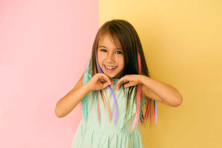 Cheerful girl with bright multicolored strands of hair laughs merrily looking at the cameras and straightening her hair with her hands. Child on a yellow-pink studio background. Stylish hair coloringの写真素材