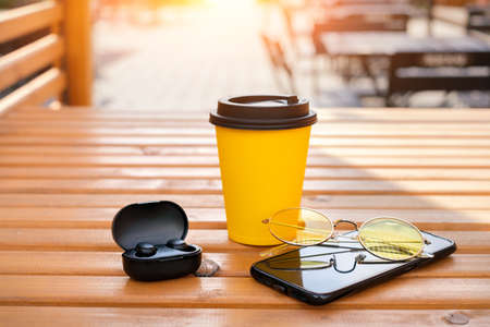 A smartphone, wireless mini earbuds in a case, a disposable cup of coffee and yellow sunglasses are lying on the table of the Gordian cafe on the terrace outside.の写真素材