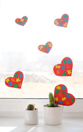 The window in the house is decorated with hearts with bright colorful puzzles. On the windowsill there are pots with various cacti and postcards for Autism Dayの写真素材