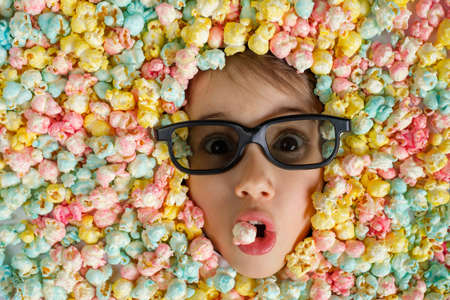 A child in 3d glasses for watching movies in the cinema with a surprised look holds popcorn in his mouth. A child's face surrounded by a pile of colorful sweet popcorn.の写真素材