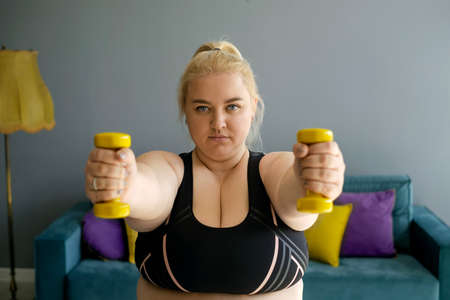A young serious plump woman trains with yellow dumbbells, the concept of doing sports at home. An...