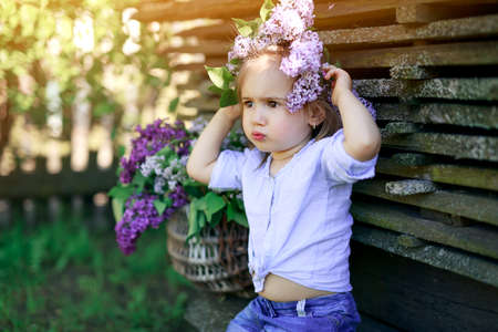Girl puts a wreath of purple lilac flowers on head with a thoughtful, dissatisfied look, seriously pouting lips. Basket with spring flowers. Baby looks pensively, dreamily, focused towards copy spaceの写真素材