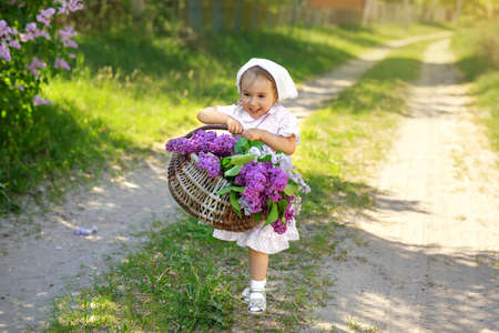 A village girl in a white dress and a kerchief carries a basket filled with spring blooming flowers and violet lilac along a country path. Funny happy pretty little child collects flowers in a bouquetの写真素材