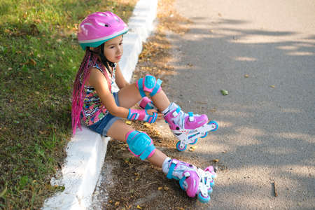 Thoughtful cute girl with Afro-pigtails and wearing sports protective gloves and a helmet sat down to rest on the sidewalk after roller skating. Kid's favorite hobby is roller skates. summer sunny dayの写真素材