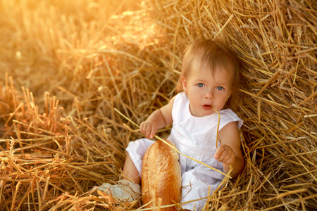 A funny cute innocent child in a white dress is sitting in a field by a haystack and holding a straw in her mouth. A hungry beautiful girl with bread. World Bread Day. children's dayの写真素材