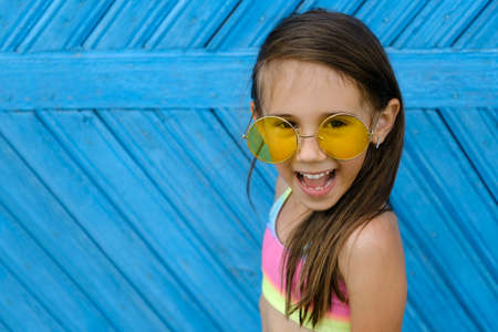 A beautiful laughing brunette girl in yellow sunglasses and a multicolored swimsuit stands against a blue wall of boards. The time of children's holidays and recreationの写真素材