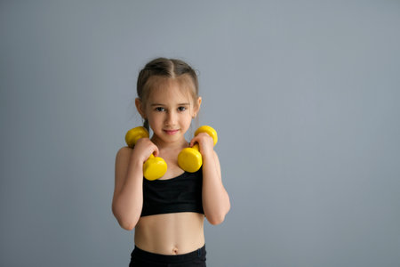 Cute beautiful sporty girl with braided pigtails plays fitness by lifting heavy dumbbells. Healthy, strong child in a sportswear. Person with sports equipment on a gray studio background copy spaceの写真素材