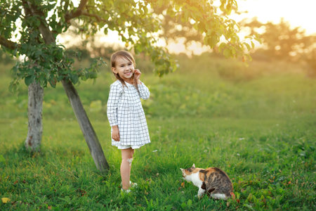 A child in the garden stands smiling sweetly next to a stray cat at sunset. Pets and their owners. Friendship of children and animals.の写真素材