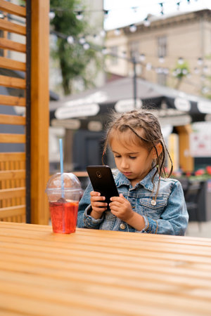 Child is sitting at table in street courtyard in cafe and looks intentionally at phone. Frequent use of smartphones and gadgets harms vision of preschool children. Girl drinks lemonade. Summer Vacation Dayの写真素材