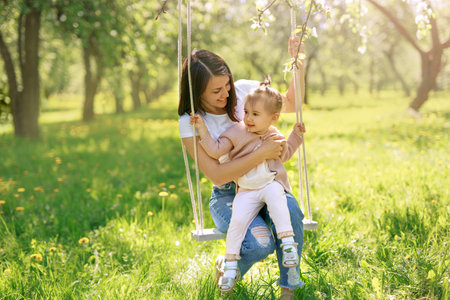 Mom and daughter swinging on a swing in a blooming garden on a hot spring day. The child has fun with his mother. mother's dayの写真素材