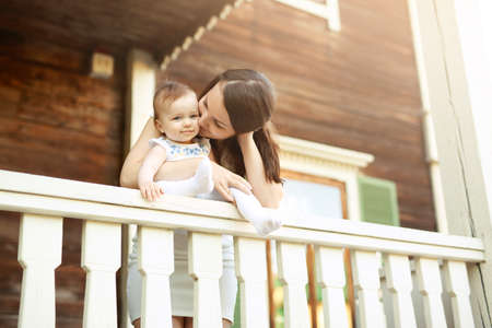 A mother hugs and kisses a little innocent baby girl on the outdoor veranda of an old wooden house. The child is sitting on the railing of the terrace. Care and upbringing of kids by parentsの写真素材