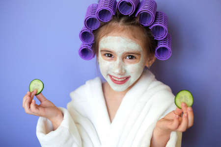 Pretty little girl brunette with curlers in hair with a clay mask on face, with slices of fresh cucumber in hands, smiling sweetly looking at the camera. Brown-eyed Lady on purple background very periの写真素材
