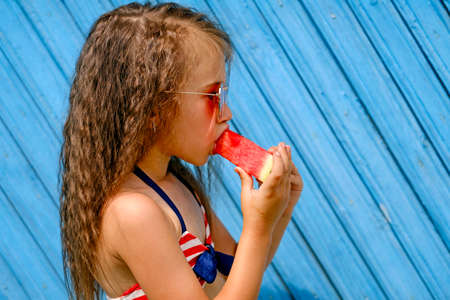 Profile of a beautiful curly-haired brunette girl biting a slice of juicy red watermelon. A child in...