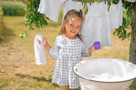 Girl holds bottle of liquid laundry detergent in hands, standing at basin for hand washing clothes. Snow-white baby underwear on clothesline. Advertising of eco-friendly hypoallergenic detergentsの写真素材