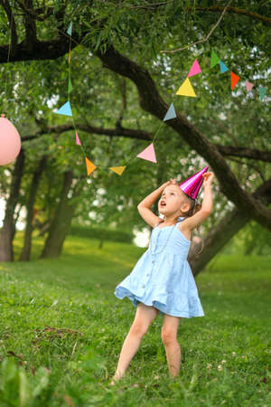 A child in a festive hat and a blue dress plays with garlands suspended from trees in the garden in honor of his birthday.の写真素材